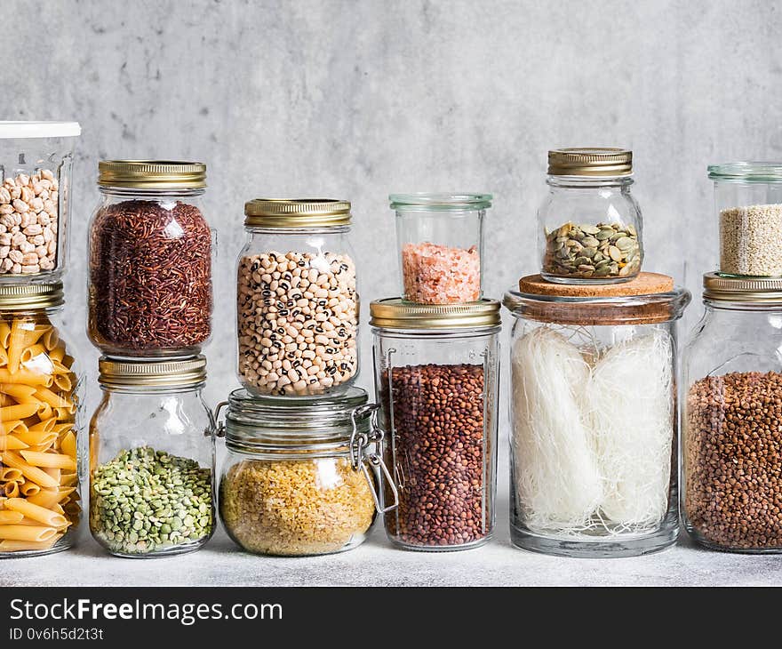 Glass jars with various cereals and seeds - peas split, sunflower and pumpkin seeds, beans, rice, pasta, oatmeal, lentils, bulgur on grey background