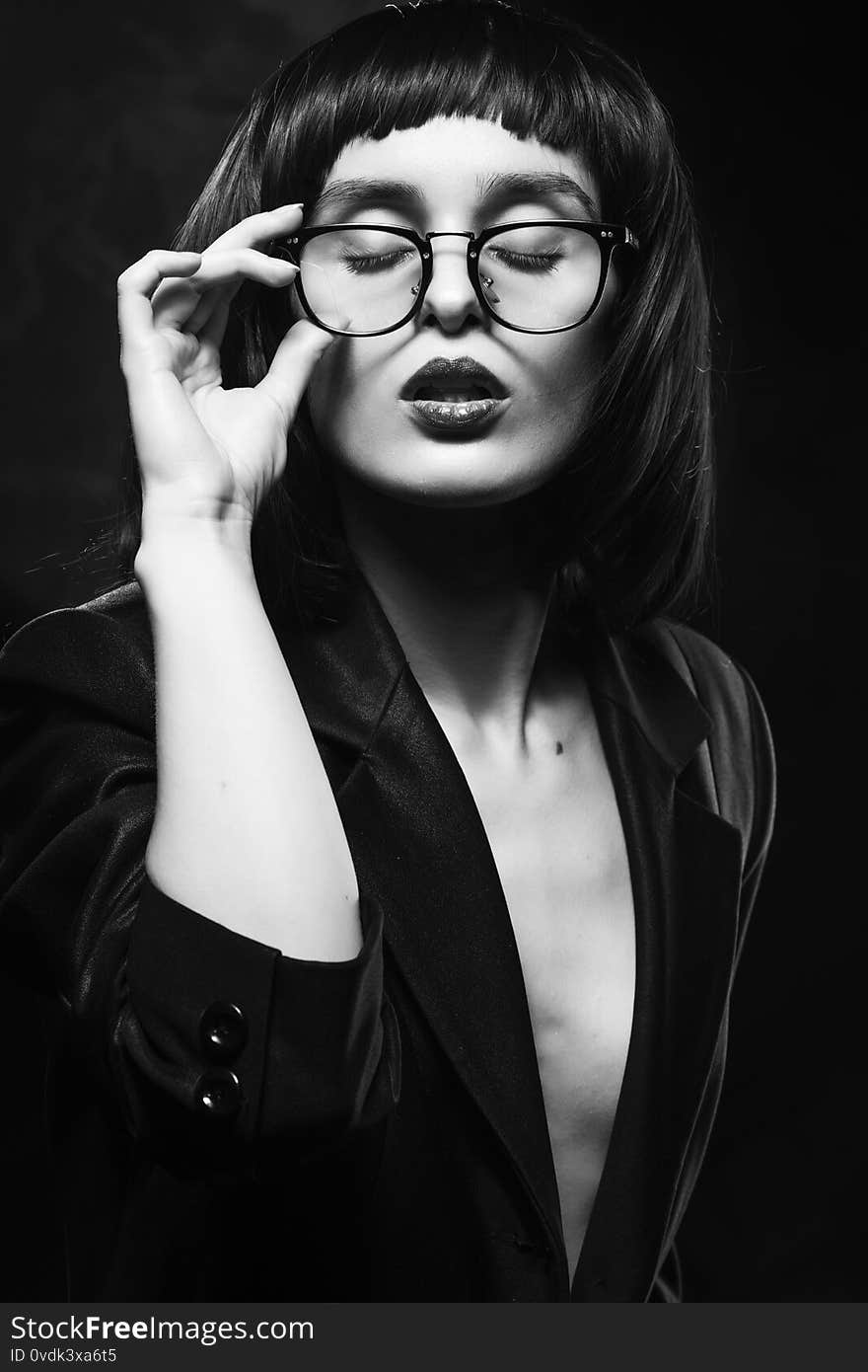 Beautiful braless slim girl with red lips, wearing a unbuttoned black blazer, glasses and brunette wig on a dark background