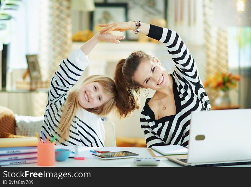 Smiling young mother and daughter in striped sweaters in home office in the modern living room in sunny day stretching. Smiling young mother and daughter in striped sweaters in home office in the modern living room in sunny day stretching