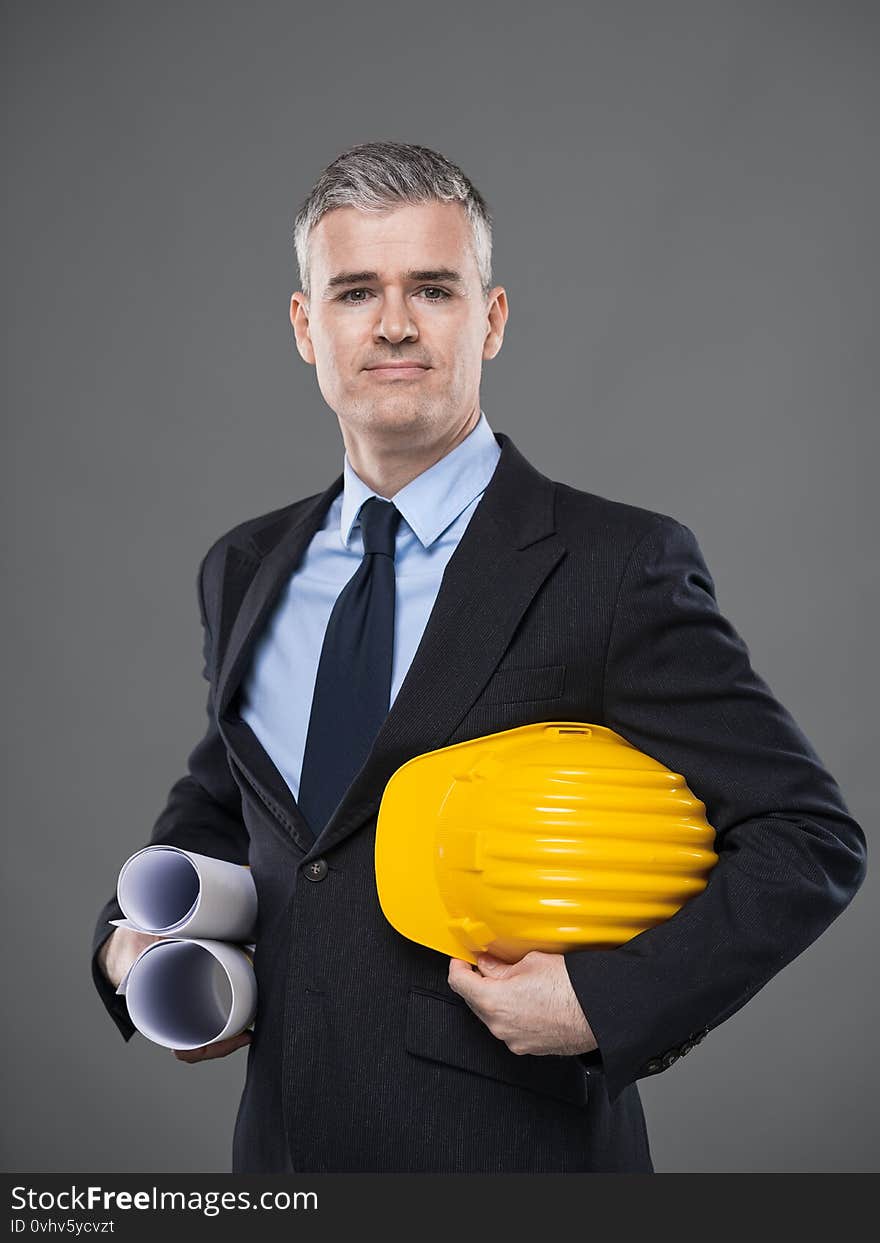 Architect or structural engineer carrying blueprints and hardhat under his arms standing smiling at the camera over grey with copy space. Architect or structural engineer carrying blueprints and hardhat under his arms standing smiling at the camera over grey with copy space