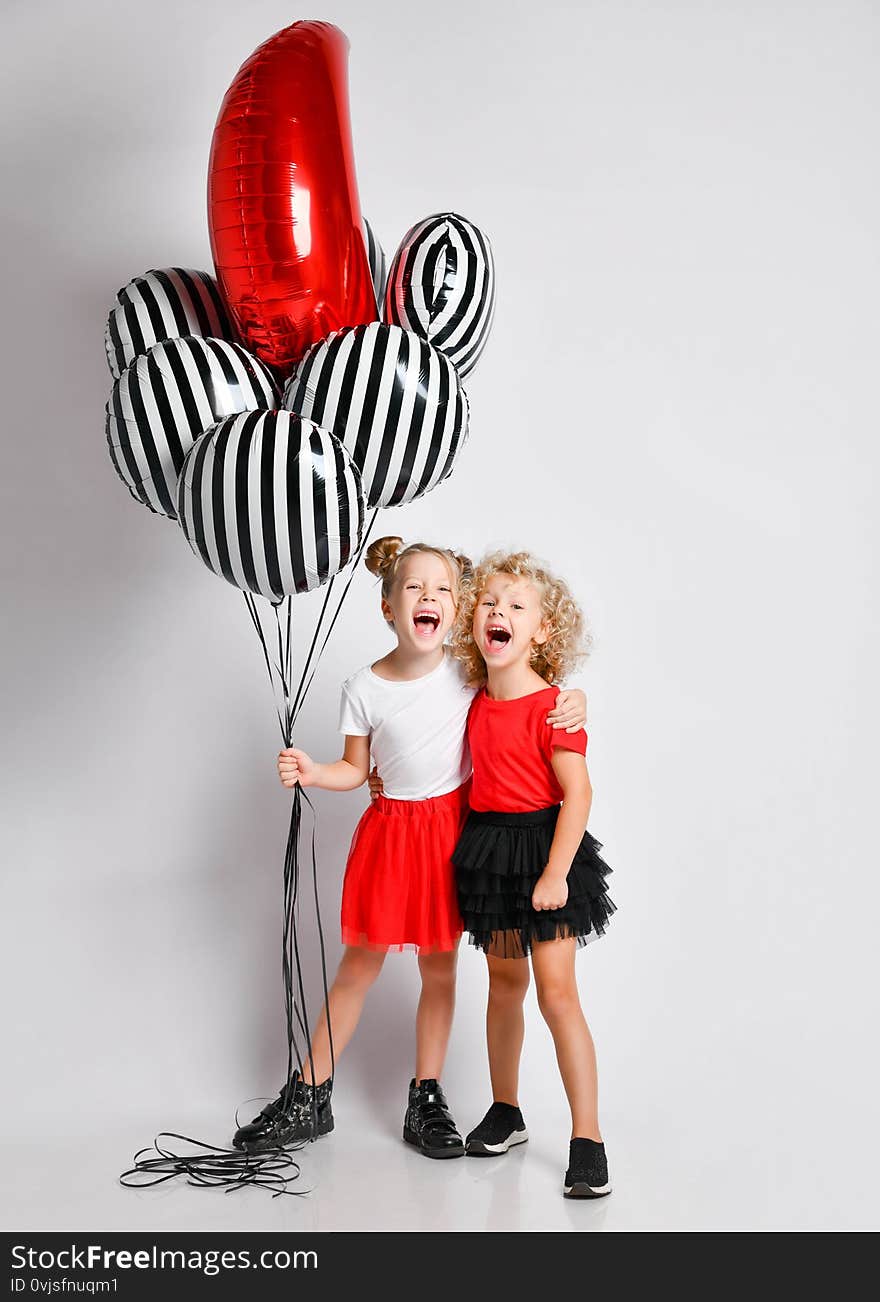 Two happy singing screaming blonde kids girls in red and black skirts and white and red t-shirts with big stylish air balloons stand together hugging. Two happy singing screaming blonde kids girls in red and black skirts and white and red t-shirts with big stylish air balloons stand together hugging