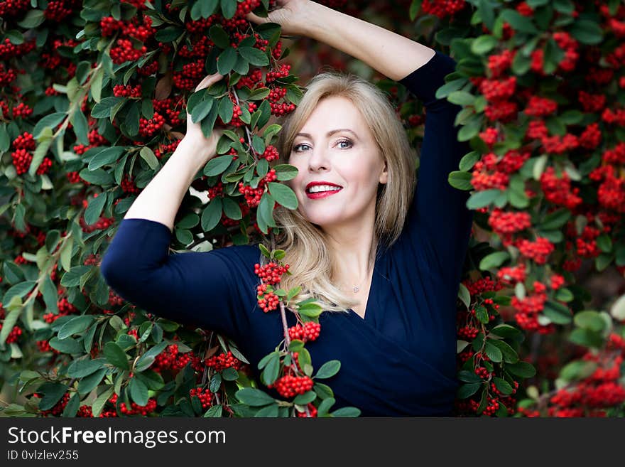 A waist up portrait of Caucasian woman with long blond hair with red near a bush with red berries.