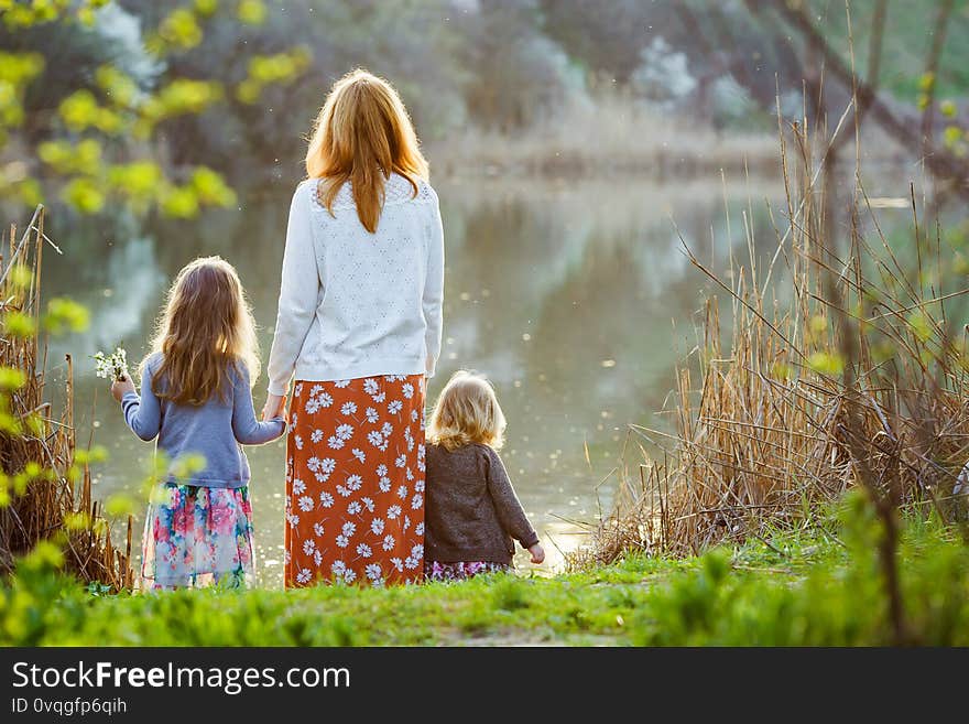 Redhead mom with two daughters standing on the banks of a small pond. spring landscape near the water. rear view. Redhead mom with two daughters standing on the banks of a small pond. spring landscape near the water. rear view.