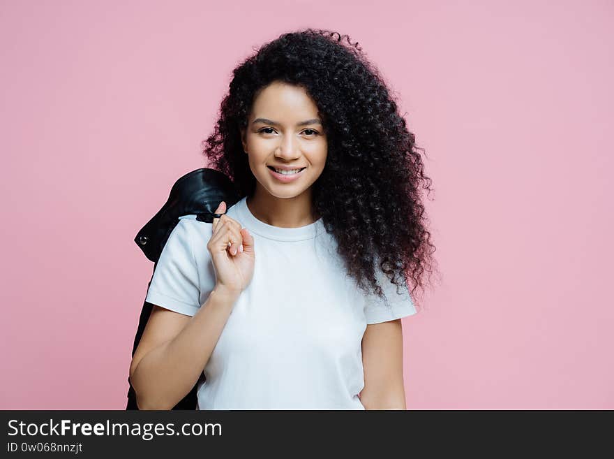 Portrait of positive ethnic woman wears white t shirt, carries jacket on shoulder, has cheerful expression, ready for picnic, enjoys day off, poses indoor against rosy background. People, lifestyle