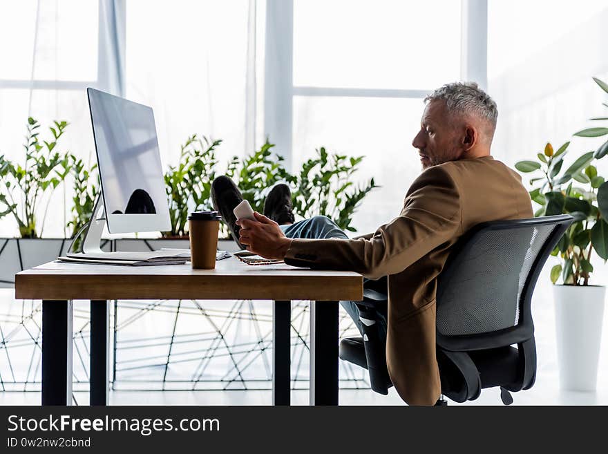 View of handsome businessman sitting near computer monitor and holding smartphone