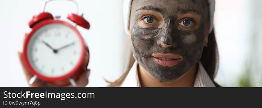 Portrait of good-looking young woman holding red clock and timing to take off facial clay mask. Lovely female looking at camera with happiness. Spa day and skincare concept. Portrait of good-looking young woman holding red clock and timing to take off facial clay mask. Lovely female looking at camera with happiness. Spa day and skincare concept