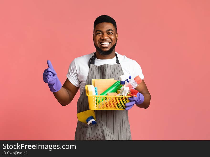 Cleaning With Pleasure. Positive Black Man Holding Container With Detergents And Household Supplies And Showing Thumb Up Over Pink Background. Cleaning With Pleasure. Positive Black Man Holding Container With Detergents And Household Supplies And Showing Thumb Up Over Pink Background