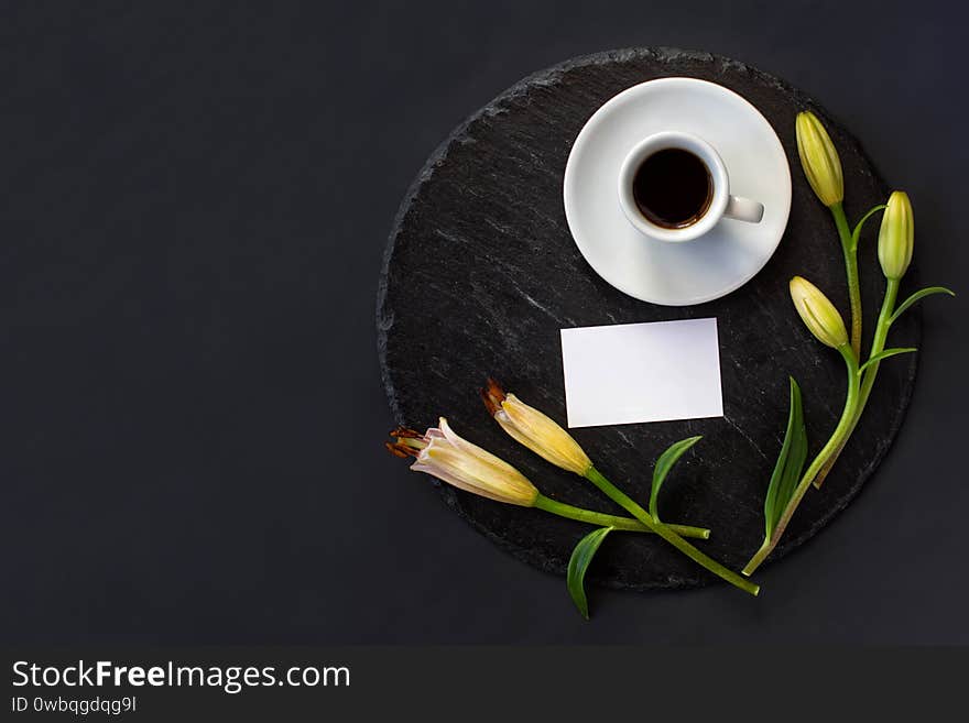 Cup of black coffee and fresh spring buds of lily flowers with clean white business card without massage on a round slate on a black background. Romantic stylish breakfast. Date on restaurant. Elegance spa. Still life flat lay. Top view. Space for text. Cup of black coffee and fresh spring buds of lily flowers with clean white business card without massage on a round slate on a black background. Romantic stylish breakfast. Date on restaurant. Elegance spa. Still life flat lay. Top view. Space for text
