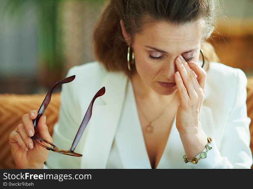 Tired elegant middle age housewife in white blouse and jacket with glasses rubbing eyes in the modern living room in sunny day. Tired elegant middle age housewife in white blouse and jacket with glasses rubbing eyes in the modern living room in sunny day