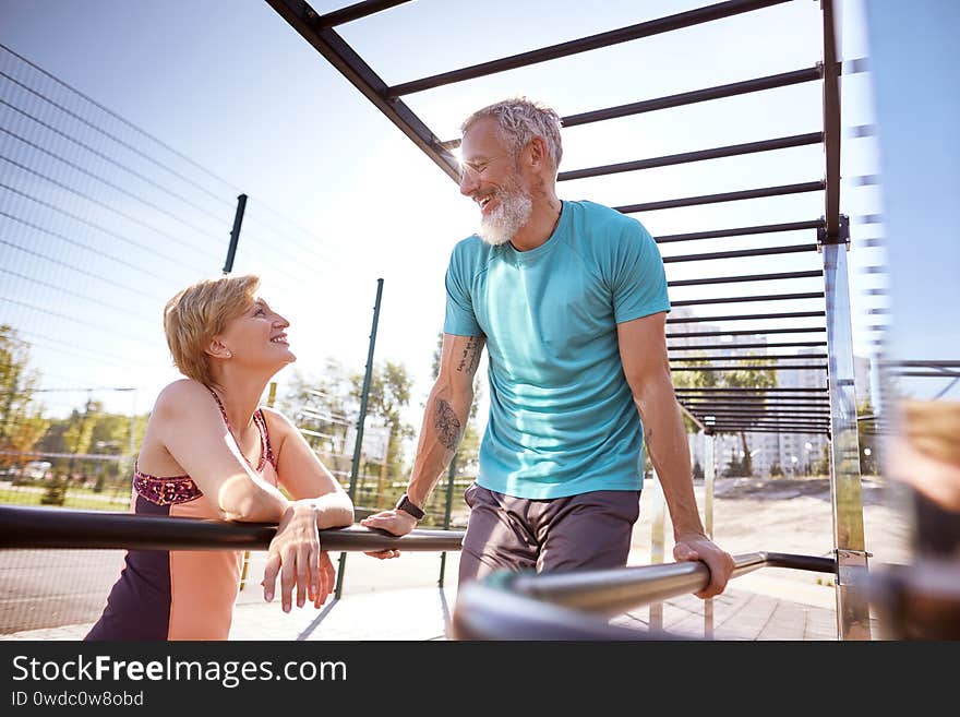 Happy mature men in a great shape doing push ups on parallel bars and talking with his smiling wife. Active healthy senior couple exercising in the morning. Fitness, sport, workout, healthy lifestyle. Happy mature men in a great shape doing push ups on parallel bars and talking with his smiling wife. Active healthy senior couple exercising in the morning. Fitness, sport, workout, healthy lifestyle