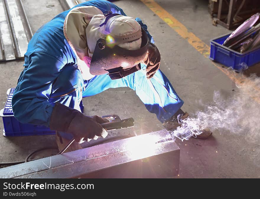 Welder is assembling the workpiece by process shielded metal arc welding SMAW. Welder in blue uniform, safety shoes, leather gloves, welding mask. He is sitting and welding. Welder is assembling the workpiece by process shielded metal arc welding SMAW. Welder in blue uniform, safety shoes, leather gloves, welding mask. He is sitting and welding