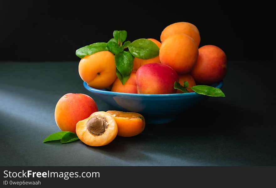 still life, ripe apricots with green foliage in a blue bowl in the rays of sunlight on a dark background