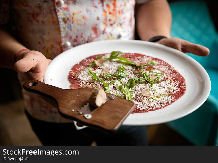 Carpaccio with parmesan, truffles and arugula on a white plate. On a wooden table. 
Chef holds in his hands