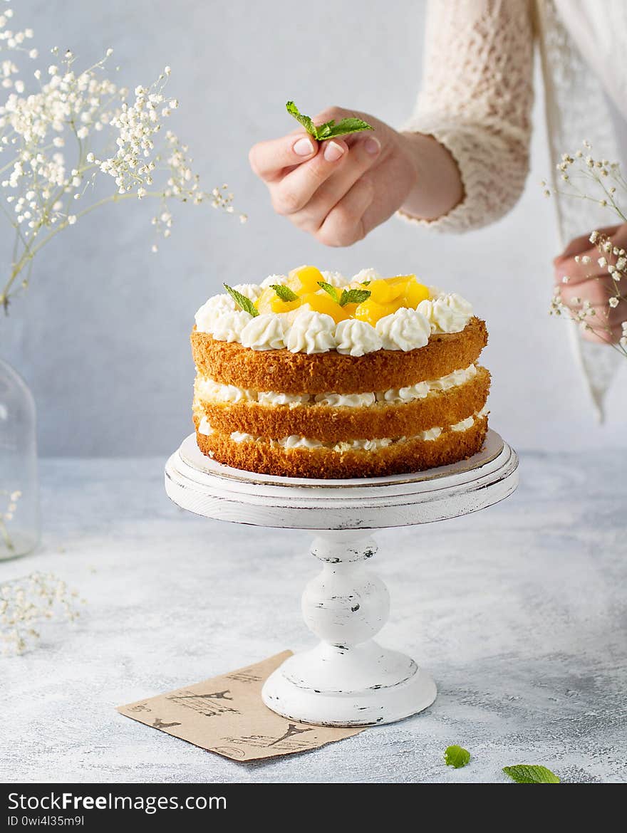 Sponge cake naked on a white stand with a leg. Female hand decorates with mint. Bright photo, white chalk flowers in the background.