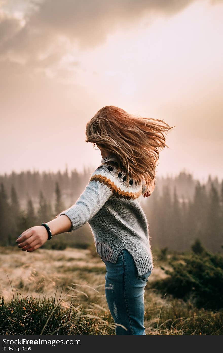 A girl stands on a mountain in a gray knitted sweater and blue jeans and looks into the distance. Wind blows hair. Dawn in the mountains. Pleasure. Peace and joy in travel