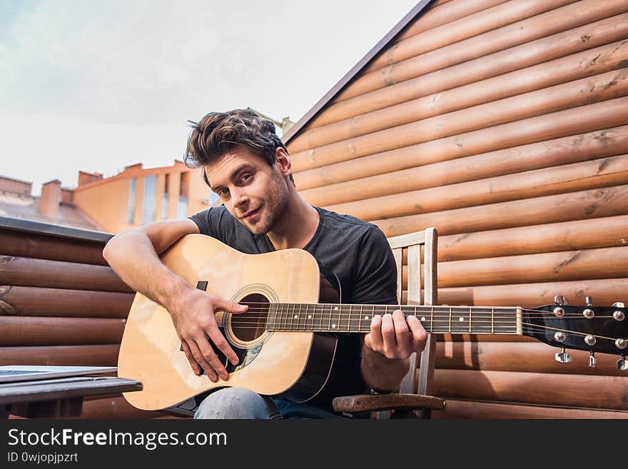 Smiling, handsome man looking at camera while sitting on balcony and playing guitar