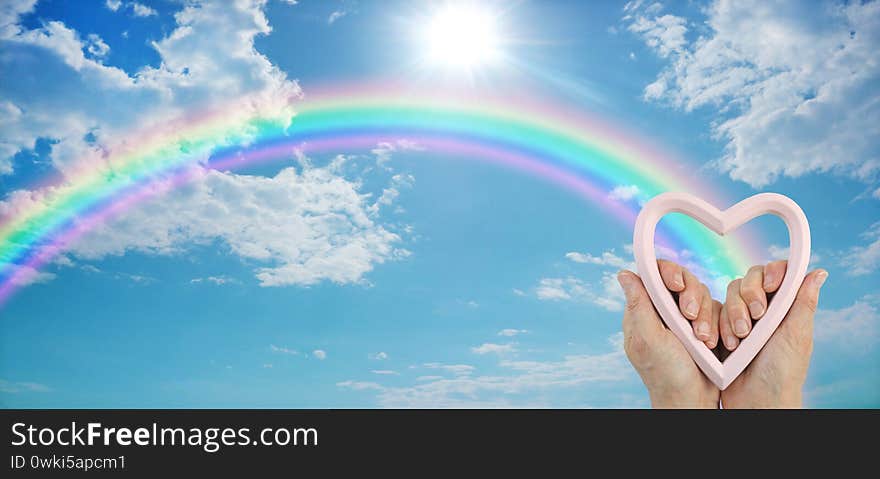 Female hands holding a heart frame with a rainbow arcing across blue sky and a sunburst above with plenty of copy space for messages. Female hands holding a heart frame with a rainbow arcing across blue sky and a sunburst above with plenty of copy space for messages