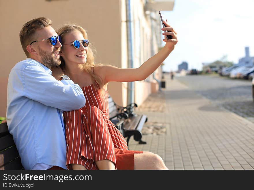 Romantic young couple in summer clothes smiling and taking selfie while sitting on bench in city street.