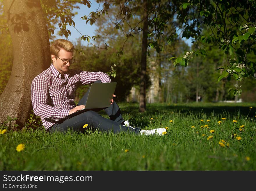 A young Caucasian man sits under a tree, typing on a laptop. Freelancer working in the park on the grass