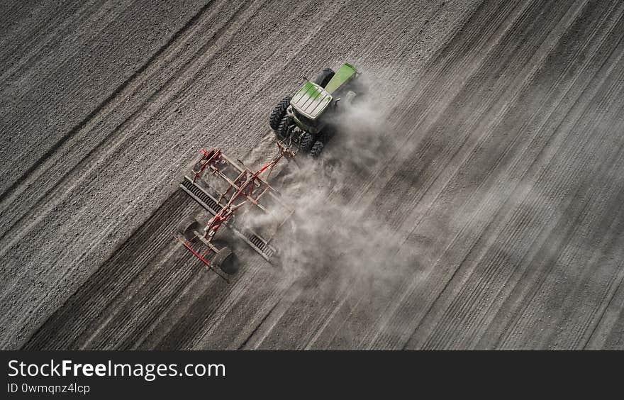 Aerial view of a clean field in which the tractor makes vertical tillage. Red tillage equipment and a green tractor on a dark gray background of the earth with drawings from the wheels. Top view. Aerial view of a clean field in which the tractor makes vertical tillage. Red tillage equipment and a green tractor on a dark gray background of the earth with drawings from the wheels. Top view