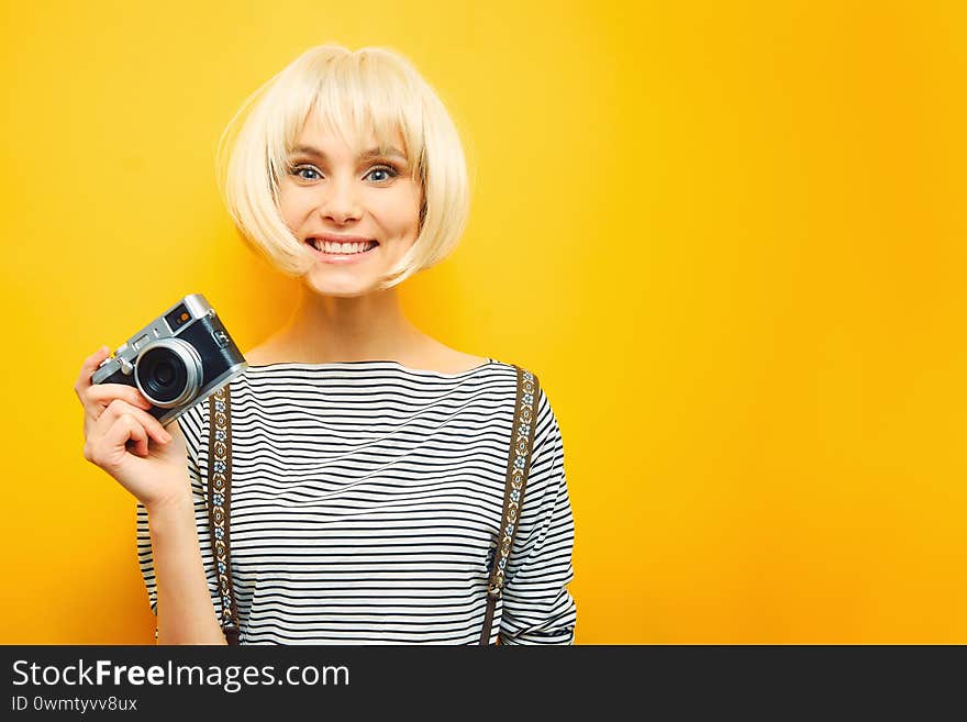 Portrait of a smilling girl with a camera in hand on a yellow background. Isolated studio. blonde wig