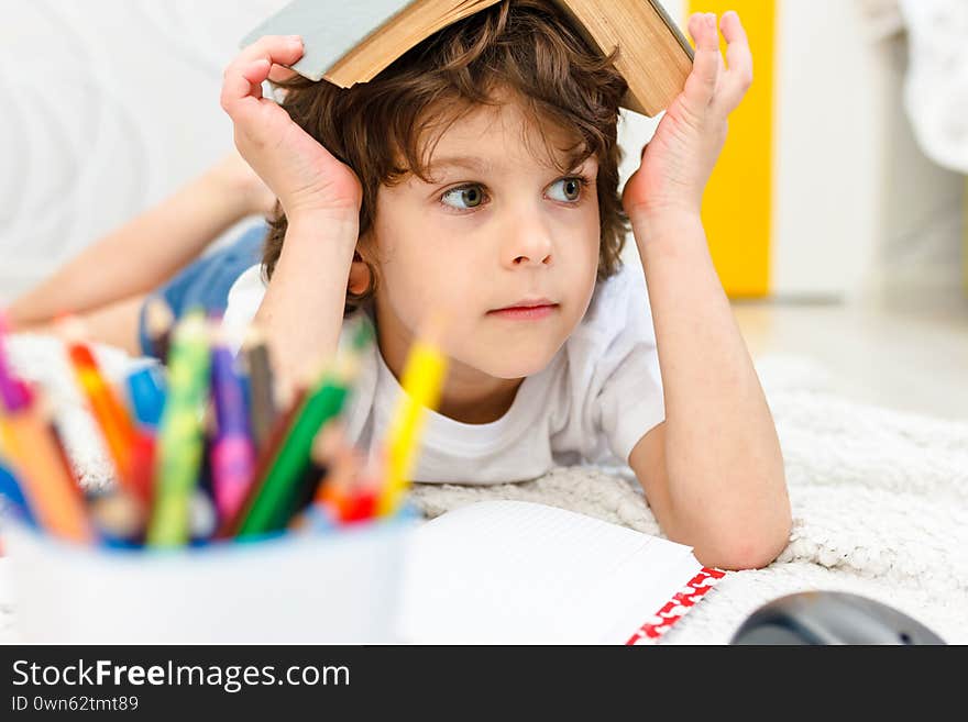 Boy does homework and holds a book on his head in light room. Concept reading, education, childhood. Social distancing and self isolation in quarantine lockdown