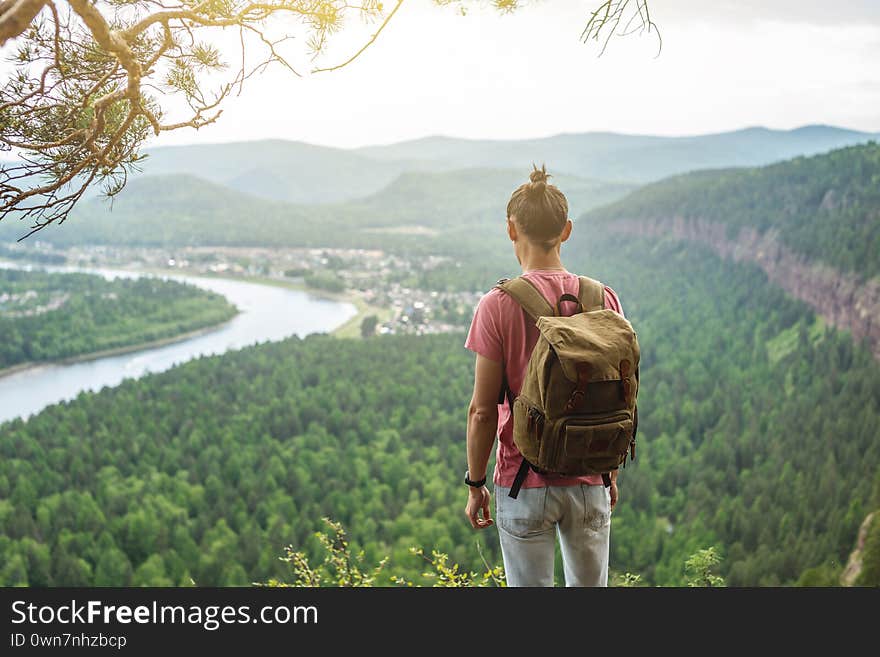 Traveler with a backpack is standing on edge of a cliff and is looking on a green valley. Freedom and unity with nature