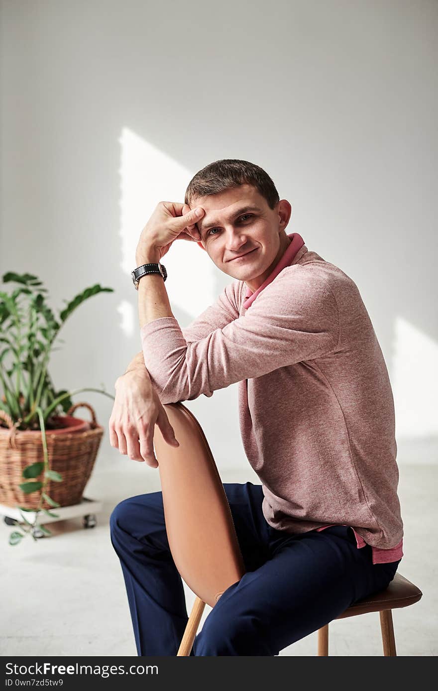 Young brunette man, wearing casual light pink jumper, sitting on chair by brick wall in light room with green plant, smiling