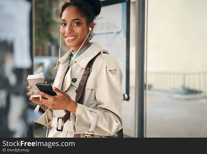 Beautiful casual African American girl in stylish trench coat with coffee to go and cellphone joyfully looking in camera at bus stop