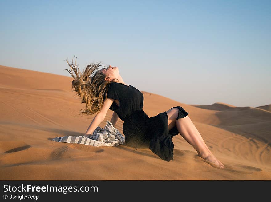 A young girl in a black dress sits on the sand in a beautiful pose with her hair developing in the wind. A young girl in a black dress sits on the sand in a beautiful pose with her hair developing in the wind