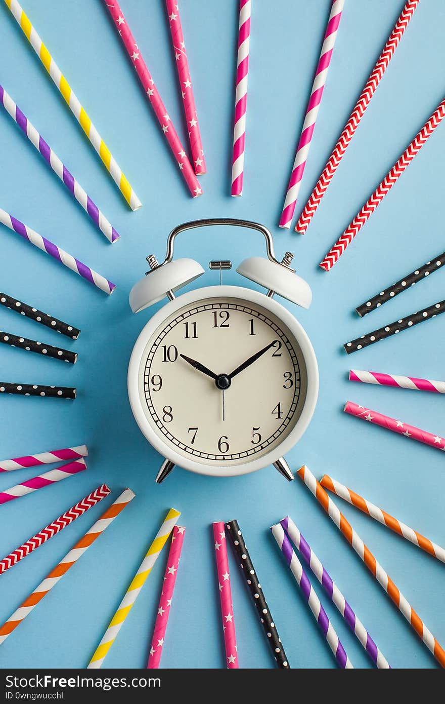 White alarm clock around multi-colored paper tubes for cocktails on a blue background. The concept of party, birthday, fun. Flatlay