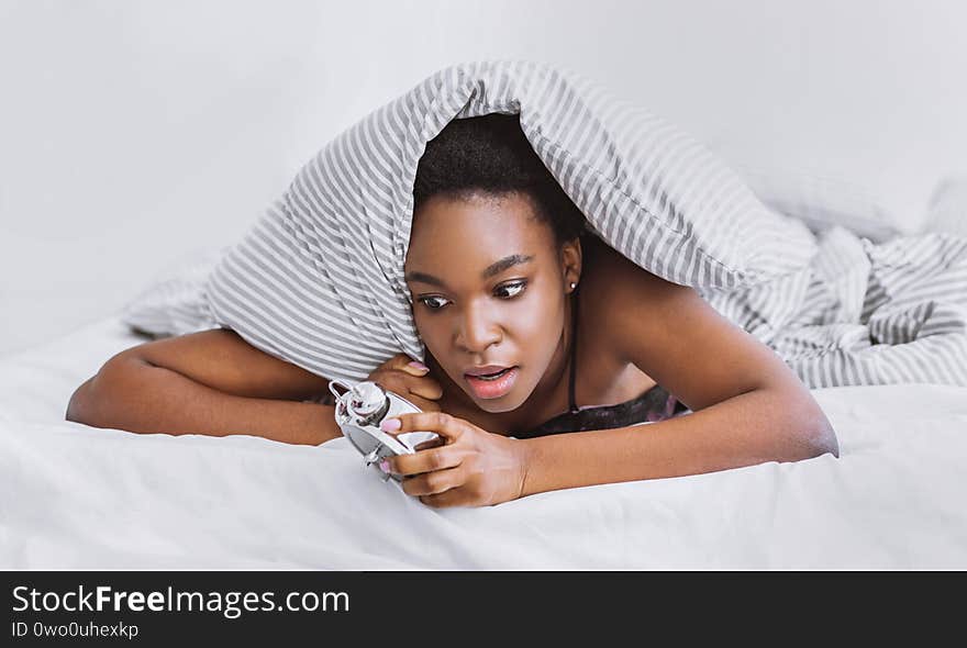 Oversleep concept. Shocked african american girl wakes up, climbs out from under covers looking at alarm clock