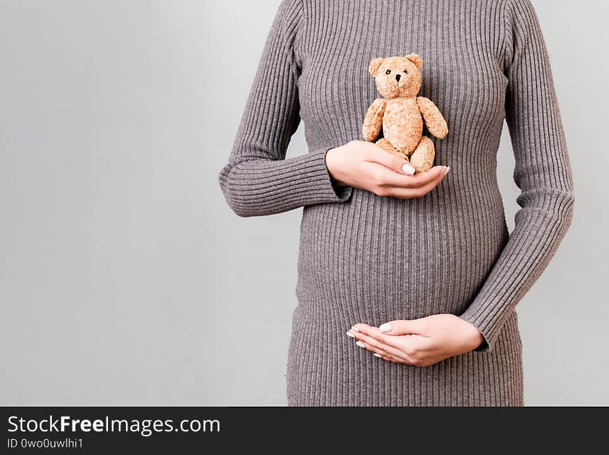 Cropped image of teddy bear in hand against pregnant woman`s belly in gray dress at gray background. Waiting for a childbirth.