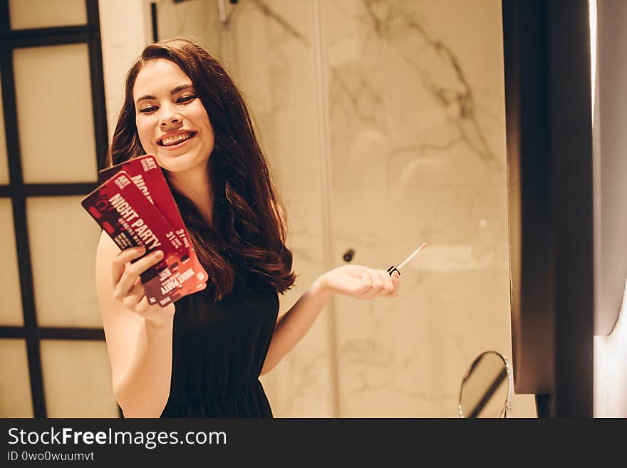 girl in black dress holding lip gloss and night party tickets in bathroom, end of quarantine concept. girl in black dress holding lip gloss and night party tickets in bathroom, end of quarantine concept