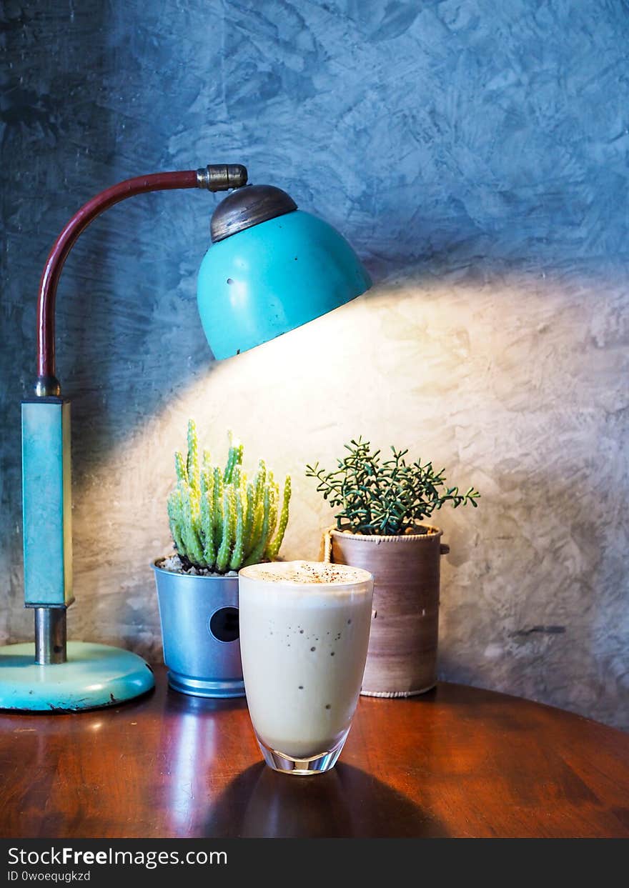 Small rest corner in house, Vintage style interior with iced coffee, old lamp and decorative trees on wooden table with gray cement loft wall. Small rest corner in house, Vintage style interior with iced coffee, old lamp and decorative trees on wooden table with gray cement loft wall