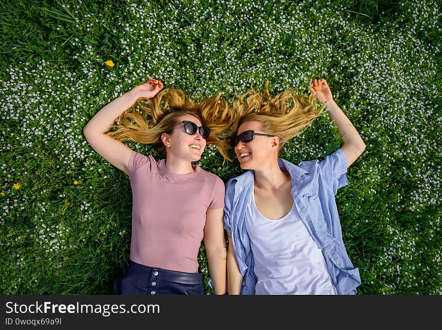 Two young happy women in a flowery meadow on sunny day, top view, close-up. Cheerful female friends lying on the lawn, looking at each other and smiling. Long hair is spread over the grass