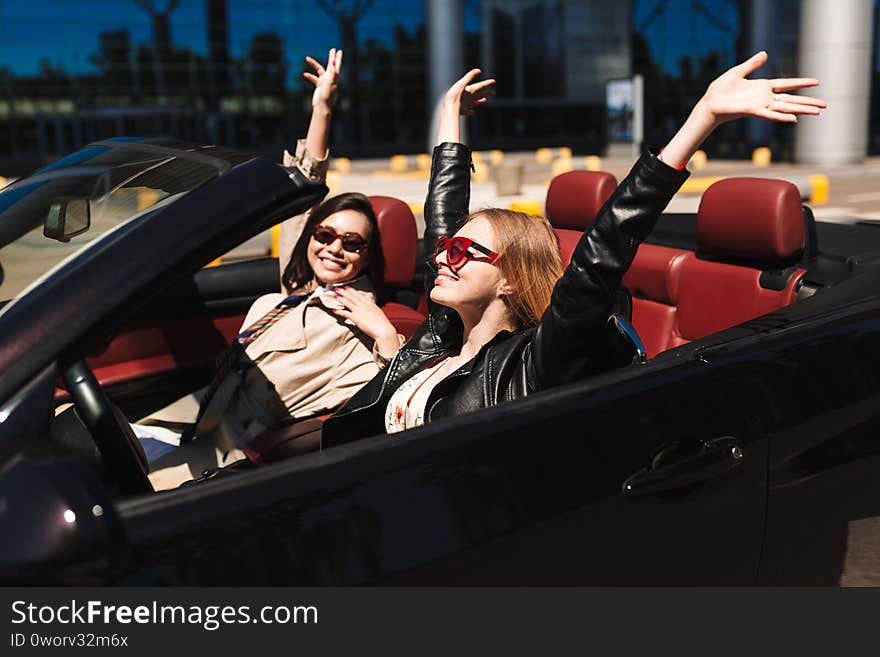 Two cheerful girls joyfully spending time together driving cabriolet car on city streets outdoor