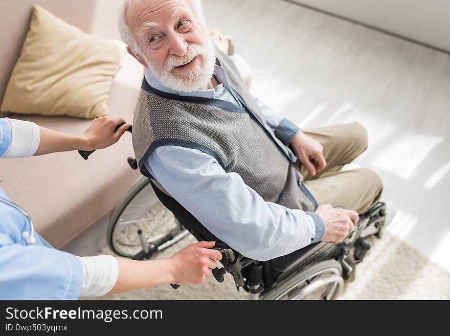 Angle view of disabled and cheerful gray haired man in wheelchair looking at nurse