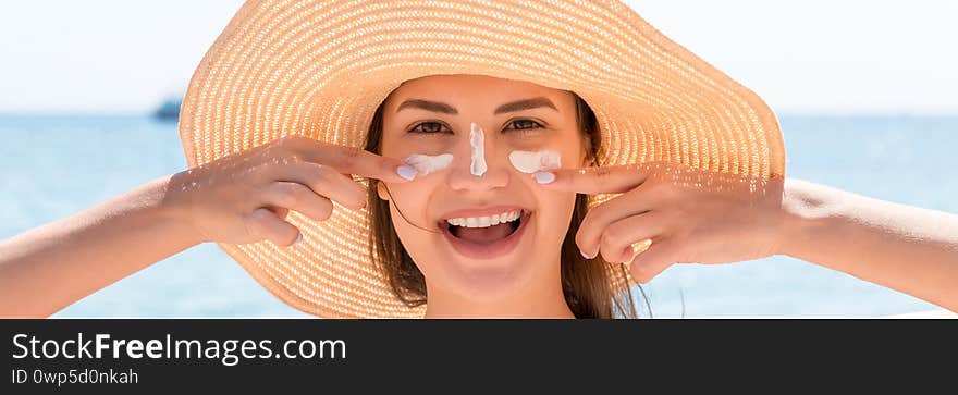 Beautiful young woman in hat is applying sunblock under her eyes and on her nose like Indian. Sun protection concept.