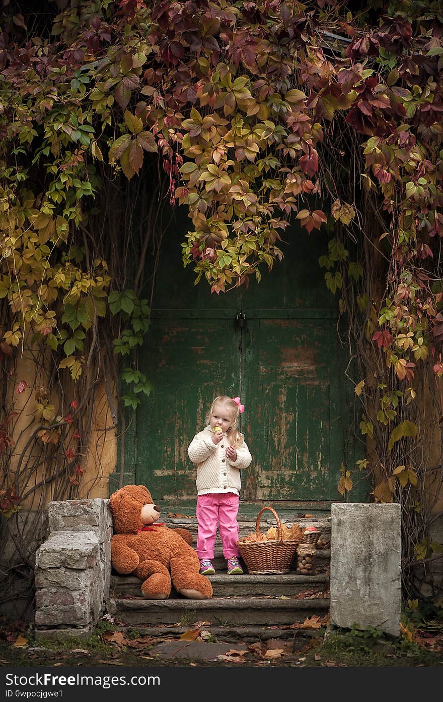 Little cute girl blonde in a light jacket and with a pink bow stands on the steps of the old porch and looks at the apple. Around leaves, on the steps lies a large teddy bear and there is a wicker basket with leaves