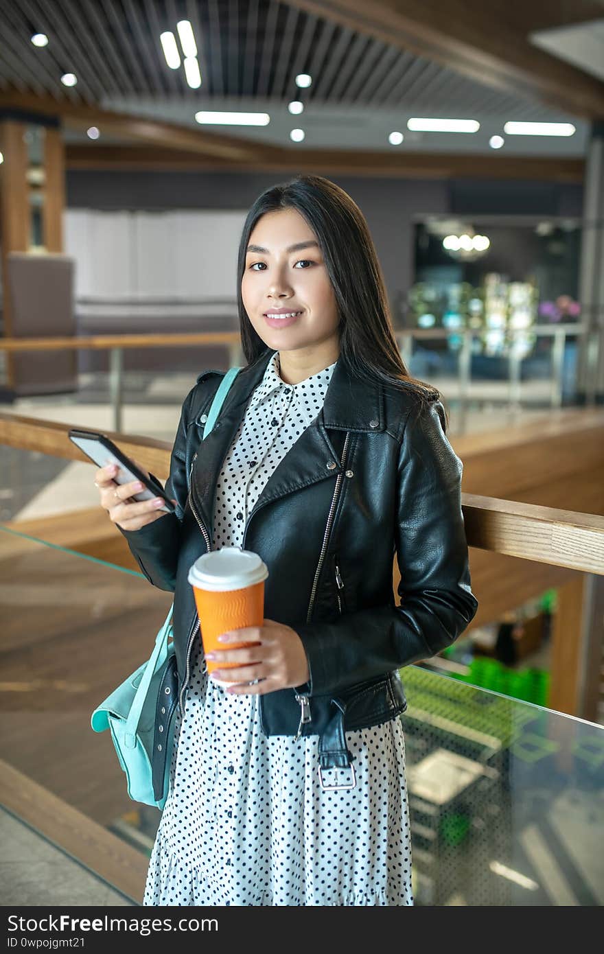 Coffee time. Young brunette female holding coffee cup and her mobile, smiling