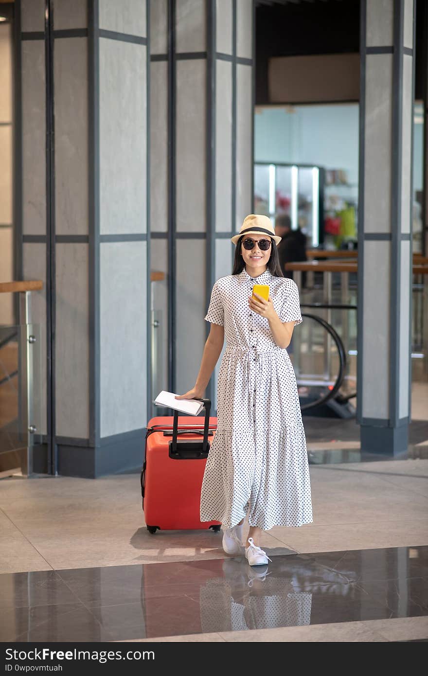 Travel time. Brunette female in straw hat and sunglasses walking with red suitcase, checking her smartphone