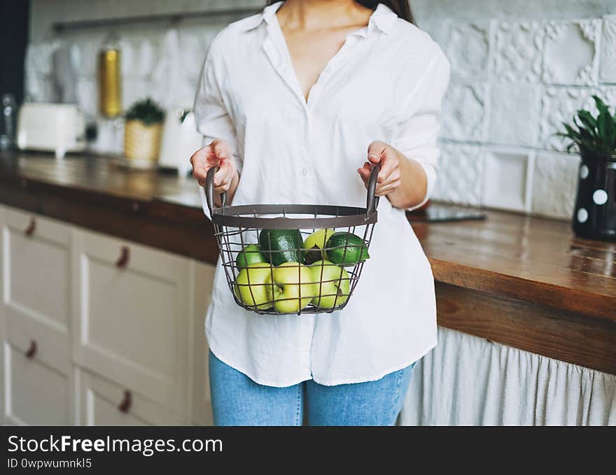 Crop photo of young woman in white shirt hold basket with green fruits and vegetables in hands on the kitchen