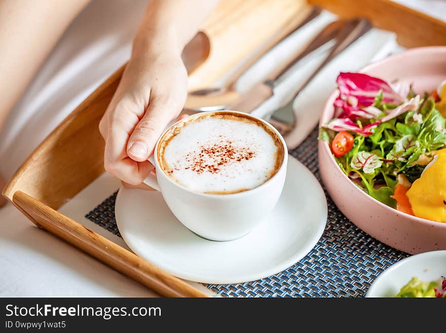 Breakfast in Bed Served with Fresh Salad and Eggs Benedict on Wooden Tray. Woman Hand Take Cup of Cappuccino Coffee. Room Service in Hotel. Breakfast in Bed Served with Fresh Salad and Eggs Benedict on Wooden Tray. Woman Hand Take Cup of Cappuccino Coffee. Room Service in Hotel