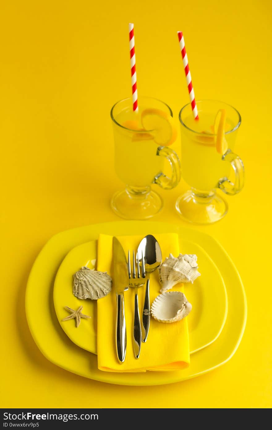 Top view of Setting stylish meal service with yellow porcelain plates,cutlery set,yellow paper napkin and two glass of lemonade on the yellow background with mussel shells.Summer collection. Top view of Setting stylish meal service with yellow porcelain plates,cutlery set,yellow paper napkin and two glass of lemonade on the yellow background with mussel shells.Summer collection.