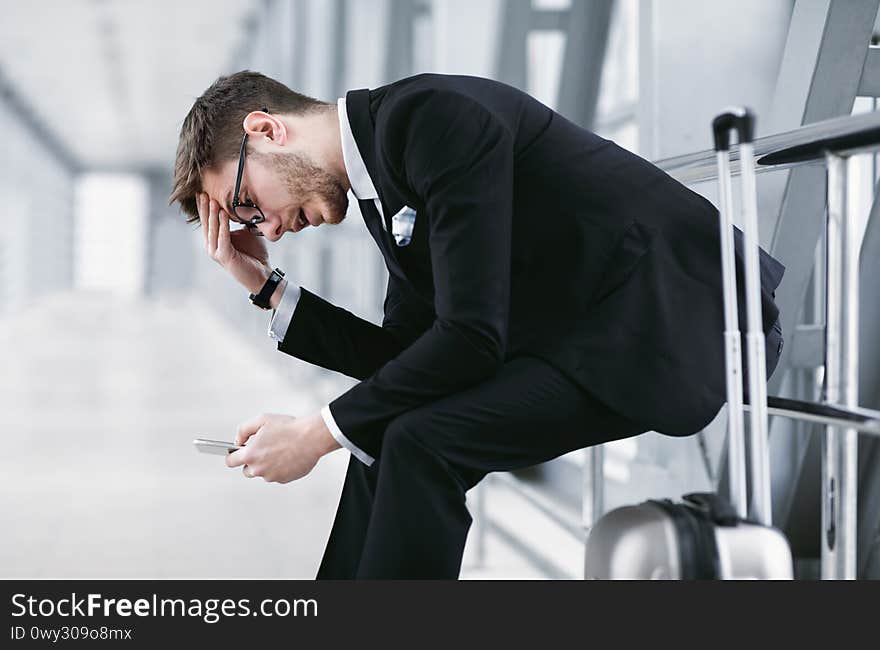 Cancelled Flight Concept. Stressed Businessman In Glasses Using Smart Phone Sitting With His Suitcase, Free Space. Cancelled Flight Concept. Stressed Businessman In Glasses Using Smart Phone Sitting With His Suitcase, Free Space