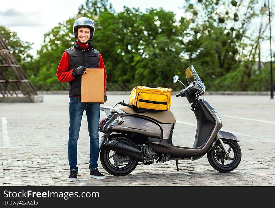 Fast Delivery. Courier Guy Standing Near Scooter Holding Paper Package Delivering Food From Restaurants Outdoors. Fast Delivery. Courier Guy Standing Near Scooter Holding Paper Package Delivering Food From Restaurants Outdoors