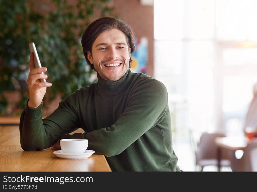 Cheerful young man holding smartphone and drinking coffee at cafe, looking at empty space