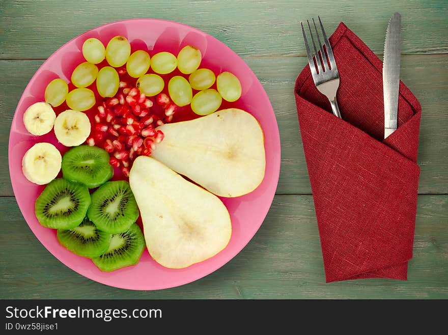 Fruit mix pear, kiwi, grapes, banana, pomegranate on a green wooden background. fruit on a pink plate with fork and knife top view. Fruit mix pear, kiwi, grapes, banana, pomegranate on a green wooden background. fruit on a pink plate with fork and knife top view