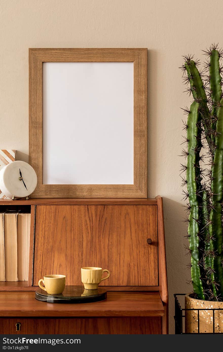 Stylish retro composition of workspace with vintage wooden cabinet, cup of coffee, design pot with cacti, mock up picture frame